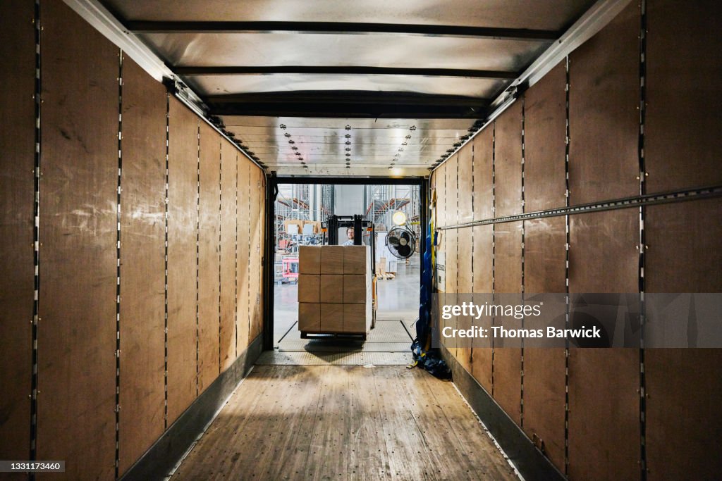 Wide view from inside of truck trailer of warehouse worker using forklift to load pallet of goods