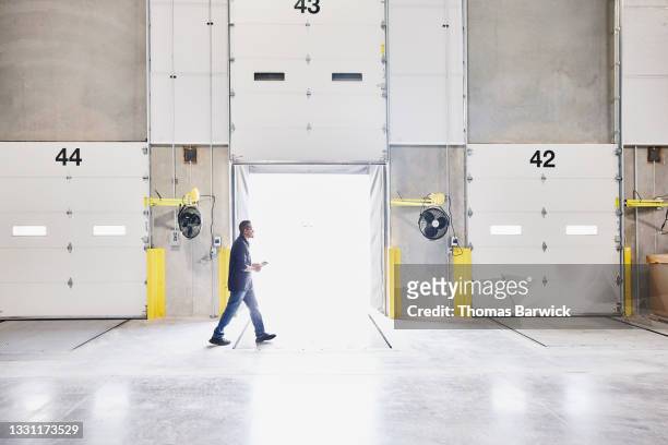 wide shot of warehouse employee holding digital tablet and walking past open loading door in warehouse - freight transportation stock-fotos und bilder