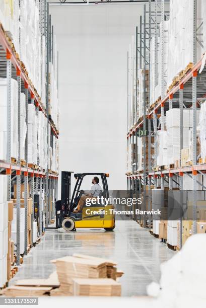 wide shot of male warehouse worker using forklift to move pallet of items in warehouse - ware house worker forklift stock pictures, royalty-free photos & images