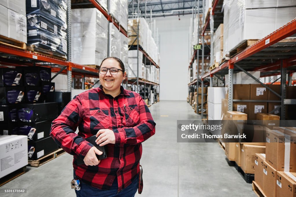 Medium view portrait of smiling female warehouse worker holding digital tablet while standing in aisle in warehouse