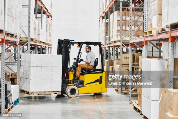 wide view of male warehouse worker driving forklift carrying pallet of goods in warehouse - carretilla elevadora fotografías e imágenes de stock