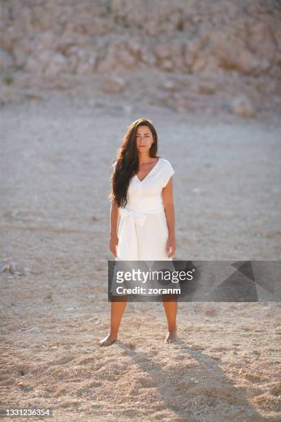 beautiful brunette in white summer dress standing barefoot on gravel in stone desert - legs apart imagens e fotografias de stock