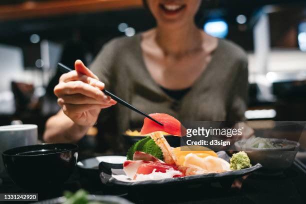 close up of smiling young asian woman enjoying delicate and fresh sashimi with chopsticks in a japanese restaurant. asian cuisine and food. eating out lifestyle - gourmet foto e immagini stock