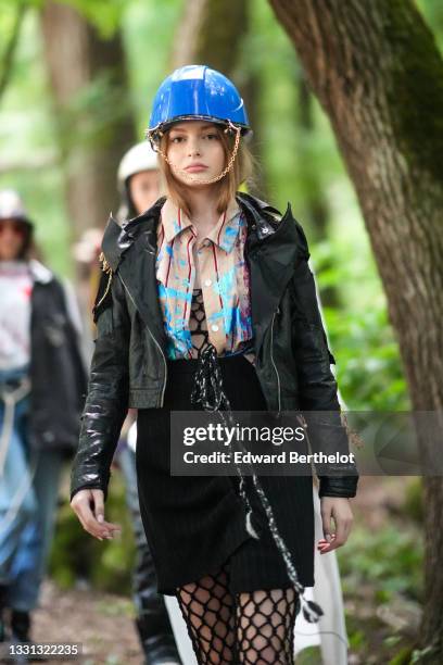 Model walks the runway and wears a royal blue construction helmet with a silver chain, a black mesh / braided jumpsuit, a beige with blue and red...