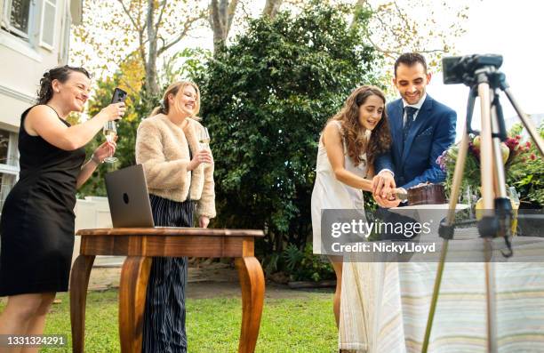 newlywed couple cutting their cake while live streaming their wedding reception - computer wedding stock pictures, royalty-free photos & images