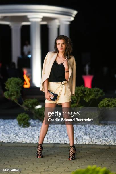 Model Rucsandra Deaconu wears a beige blazer jacket, a black sleeveless tank top with embroidery, white shorts, a clutch, lace-up high heels shoes,...