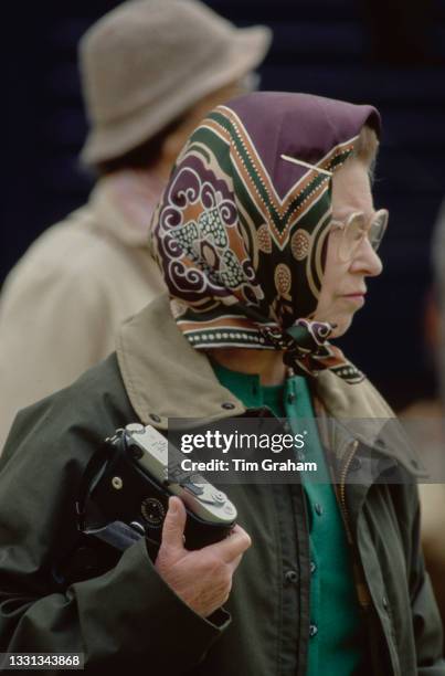 British Royal Queen Elizabeth II, wearing a headscarf and a waxed jacket, and holding a Leica M6 camera, attends the Royal Windsor Horse Show, held...