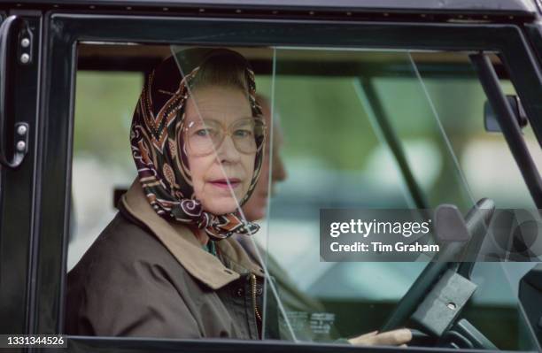 British Royal Queen Elizabeth II, wearing a headscarf and a waxed jacket, driving a Land Rover Defender at the Royal Windsor Horse Show, held at...