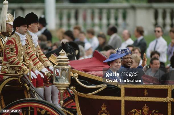 British Royal Queen Elizabeth II, wearing a blue outfit with a matching hat, sits alongside Ibrahim Babangida during the carriage procession, part of...