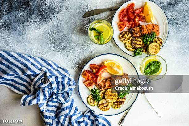 two servings of grilled salmon steak with roasted courgettes, tomatoes, lemon and a glass of lemon water - ketogenic diet stock pictures, royalty-free photos & images