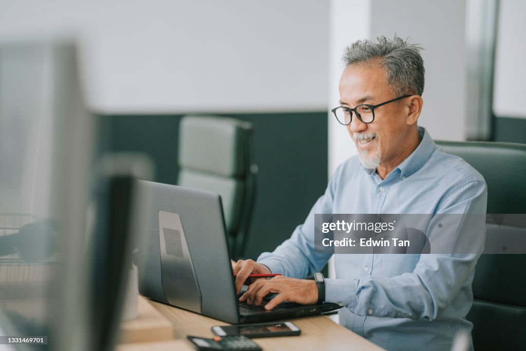 Asian chinese senior man with facial hair using laptop typing working in office open plan