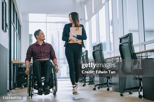 Indian white collar male worker in wheelchair having cheerful discussion conversation with his female asain chinese colleague coworking at walkway corridor Indian white collar male worker in wheelchair having cheerful discussion conversation with his female asain chinese colleague coworking at walkway corridor