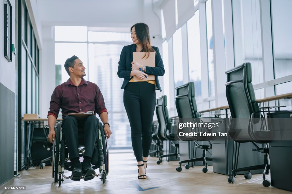 Indian white collar male worker in wheelchair having cheerful discussion conversation with his female asain chinese colleague coworking at walkway corridor