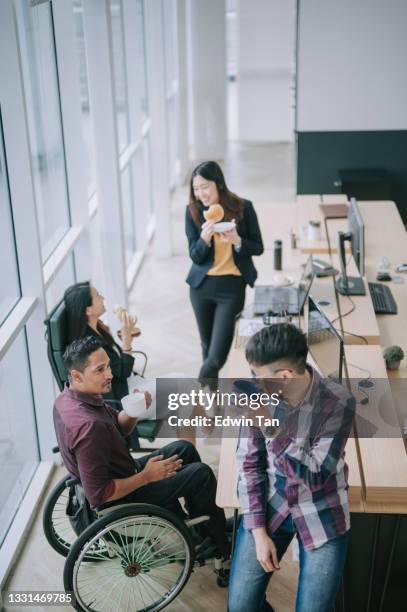 high angle view asian colleague with indian coworker in wheelchair taking a break eating sharing bread snack at their office workstation - acesso para deficientes imagens e fotografias de stock
