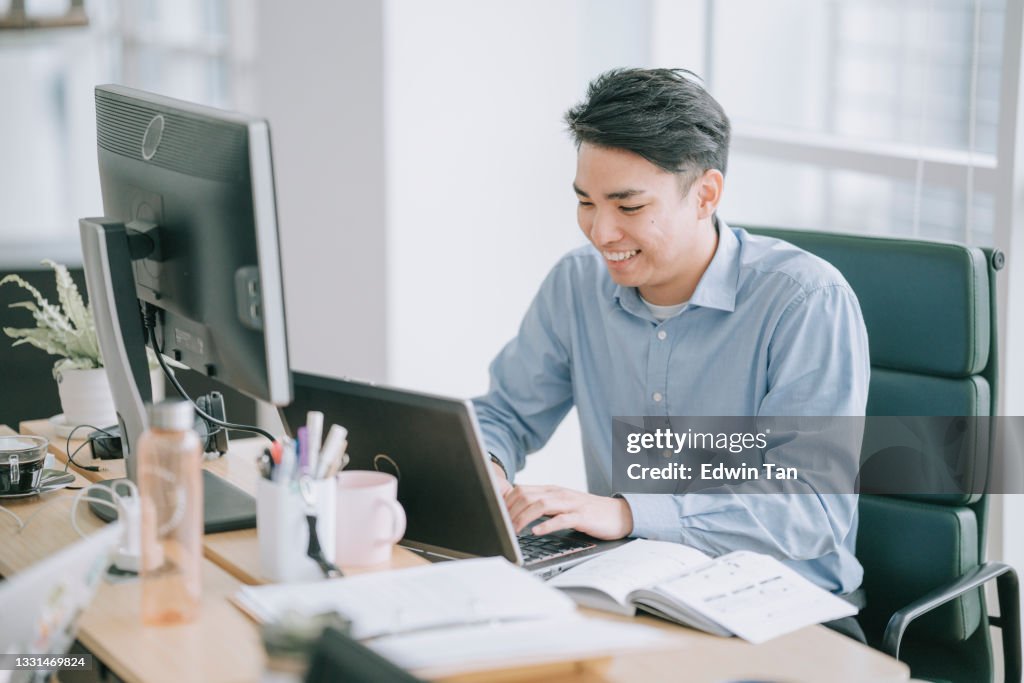Asian chinese man typing in front of laptop with multiple screen at his work place