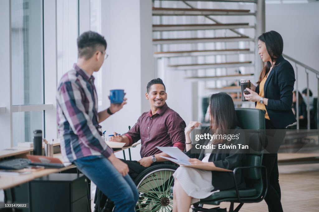 Indian white collar male worker in wheelchair having cheerful discussion leading conversation with colleague in creative office workstation beside window