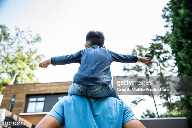 young boy riding on his father's shoulders in front of modern home - schouder stockfoto's en -beelden