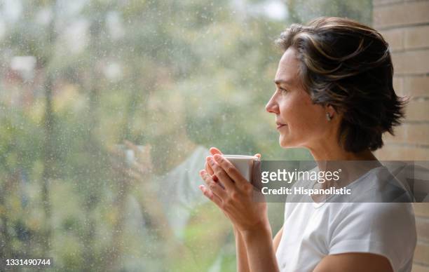 donna che beve una tazza di caffè mentre guarda fuori dalla finestra - donne mature foto e immagini stock