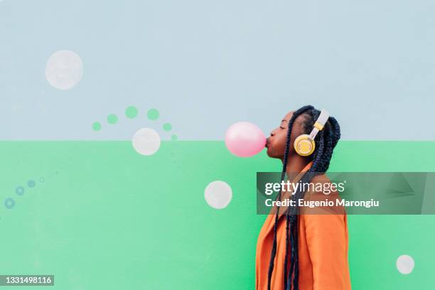 italy, milan, stylish woman with headphones blowing gum against wall - abrigo de colores fotografías e imágenes de stock
