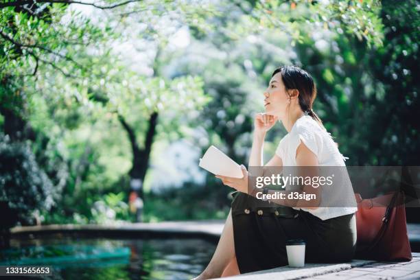 young asian woman sitting by the pond in park, in thought while reading a book. having a relaxing time enjoying the sunny day outdoors in the city. enjoying a technology-free moment - mujer leyendo libro en el parque fotografías e imágenes de stock