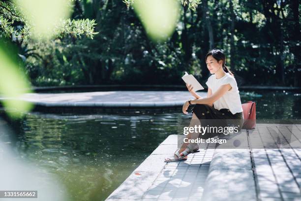 young asian woman sitting by the pond in park, reading a book and having a relaxing time enjoying the sunny day outdoors with coffee in the city. enjoying a technology-free moment - mujer leyendo libro en el parque fotografías e imágenes de stock