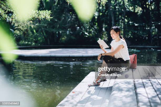 young asian woman sitting by the pond in park, reading a book and having a relaxing time enjoying the sunny day outdoors with coffee in the city. enjoying a technology-free moment - escapism reading stock pictures, royalty-free photos & images