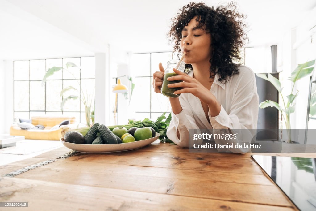 Young african american woman drinking green juice with reusable bamboo straw in loft apartment. Copy space