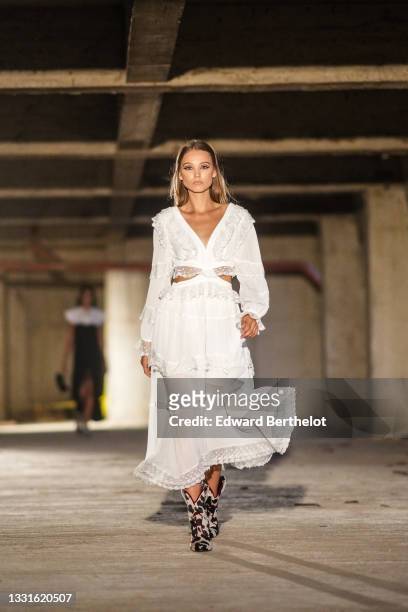 Model walks the runway and wears a white V-neck / long puffy sleeves / cut-out / ruffled / embroidered lace details / flowing long dress, black and...