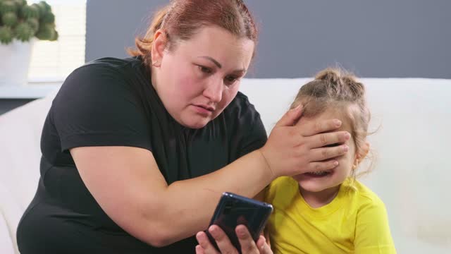 Plus size woman showing to her little daughter information on the smartphone and suddenly mother covers her daughter eyes, so that she does not see bad thinks.