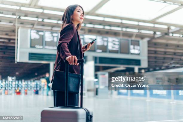 young businesswoman holding phone and carrying suitcase in airport - airport luggage fotografías e imágenes de stock