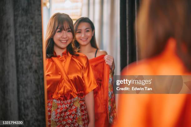 sposa cinese asiatica che prova abiti da sposa tradizionali cinesi guardando lo specchio con l'aiuto di sua sorella al negozio nuziale - chinese wedding dress foto e immagini stock