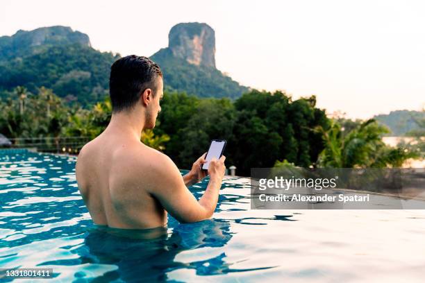 young man using smartphone in the swimming pool surrounded by tropical mountains - majestic hotel stock pictures, royalty-free photos & images