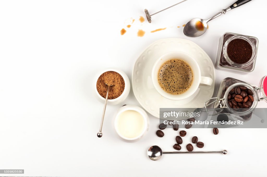 High angle view of coffee cup, roasted coffee beans, spoon, milk cream, raw cane sugarand ground coffee on white background.