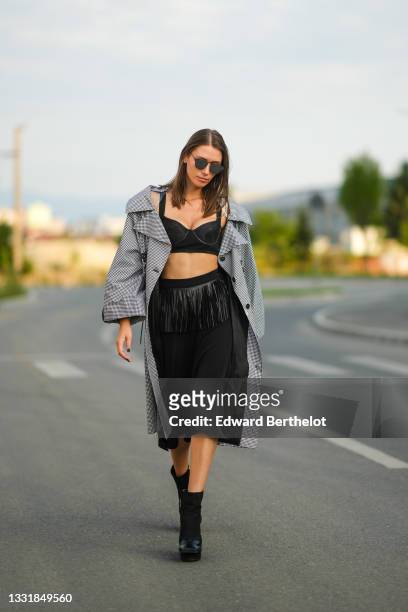 Landiana Cerciu wears sunglasses, a black and white checked pattern long trench coat, black cropped top / bras / bralette, a black midi skirt with...