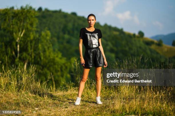 Landiana Cerciu wears a black and white t-shirt with a printed picture and the slogan "Tres Jolie", black leather mini shorts, white sneakers, on...