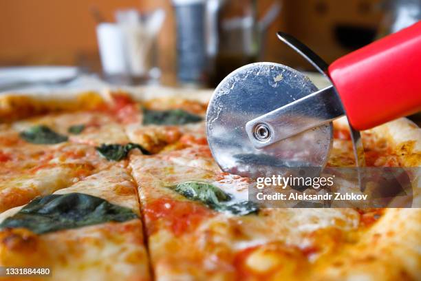 a circle of pizza margarita with mozzarella, basil leaves and sauce on a wooden stand or cutting board, on the table in a pizzeria, cafe or fast food restaurant. next to the knife for slicing pizza. traditional italian cuisine. vegetarian food. close-up. - ピザカッター ストックフォトと画像