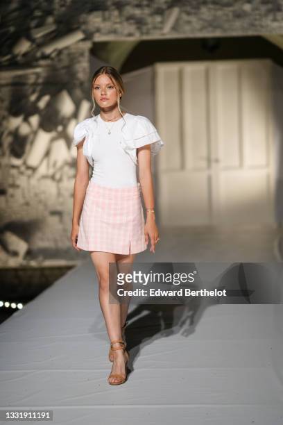 Model walks the runway and wears gold pendant earrings, a silver pendant chain necklace, a white ribbed t-shirt with short puffy / ruffled sleeves, a...
