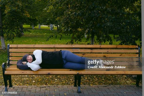 an exhausted, tired, lonely traveler or tourist is resting and sleeping on a wooden bench in the park on an autumn day. a homeless man or a young unemployed guy is lying on a bench with a backpack under his head. - homeless person stockfoto's en -beelden
