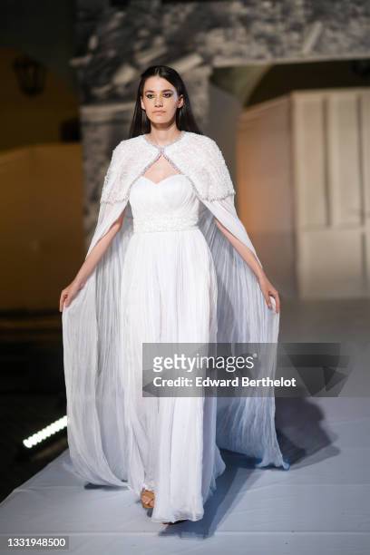 Model walks the runway and wears a white embroidered sequins shoulder long tulle train, a white off-shoulders / sweetheart long flowing / pleated /...