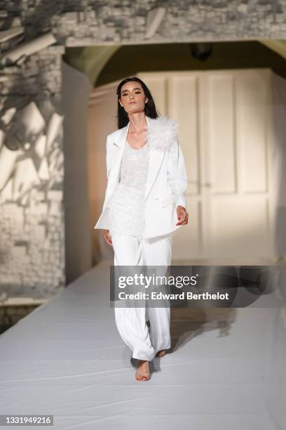 Model walks the runway and wears a white blazer jacket with white feathers on left shoulder, a white V-neck long t-shirt with white embroidered...