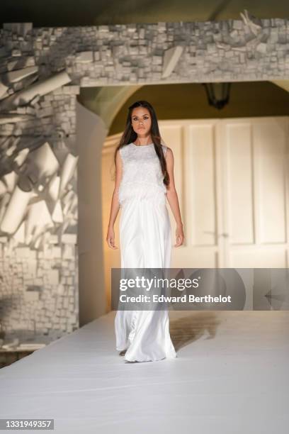 Model walks the runway and wears a white with embroidered flower sequins pattern and feathers tank-top, a white shiny satin flowing long skirt,...