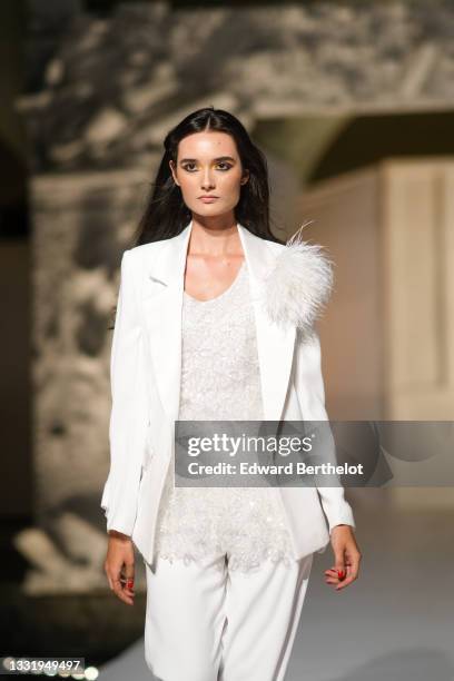 Model walks the runway and wears a white blazer jacket with white feathers on left shoulder, a white V-neck long t-shirt with white embroidered...