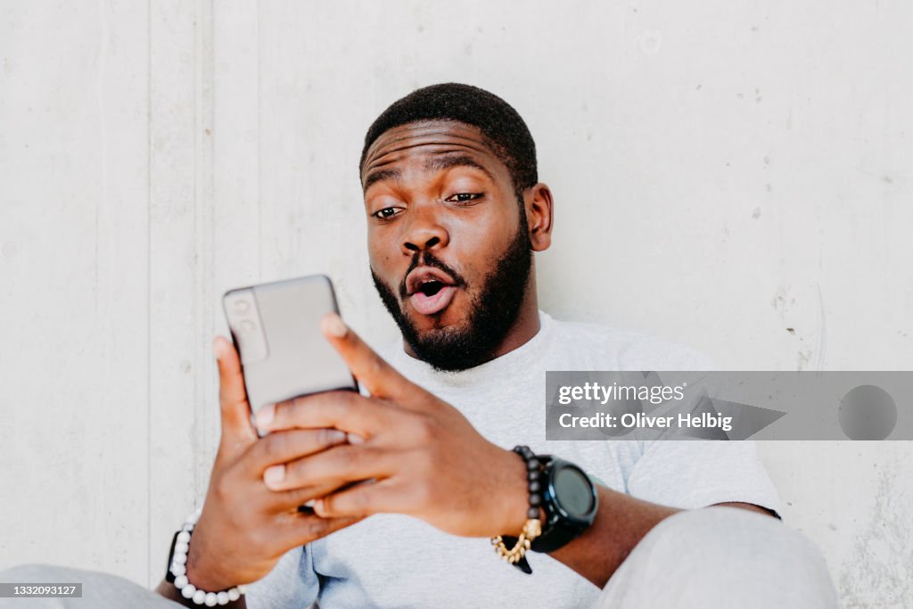 Young african american man with surprised facial expression hold smartphone in his hands, typing message, chatting with friends in social networks, playing online game, isolated against grey wall