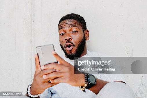 Young african american man with surprised facial expression hold smartphone in his hands, typing message, chatting with friends in social networks, playing online game, isolated against grey wall Young african american man with surprised facial expression hold smartphone in his hands, typing message, chatting with friends in social networks, playing online game, isolated against grey wall