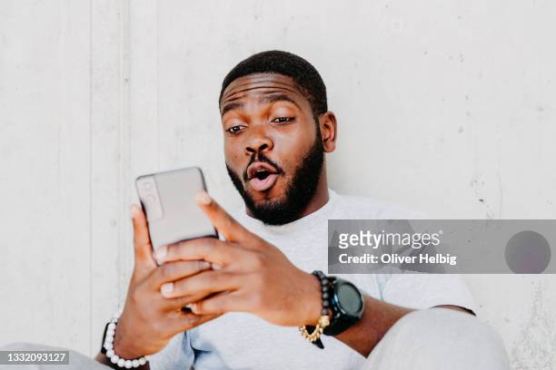 young african american  man with surprised facial expression hold smartphone in his hands, typing message, chatting with friends in social networks, playing online game, isolated against grey wall - eccitazione foto e immagini stock