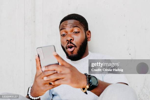 young african american  man with surprised facial expression hold smartphone in his hands, typing message, chatting with friends in social networks, playing online game, isolated against grey wall - ado imagens e fotografias de stock