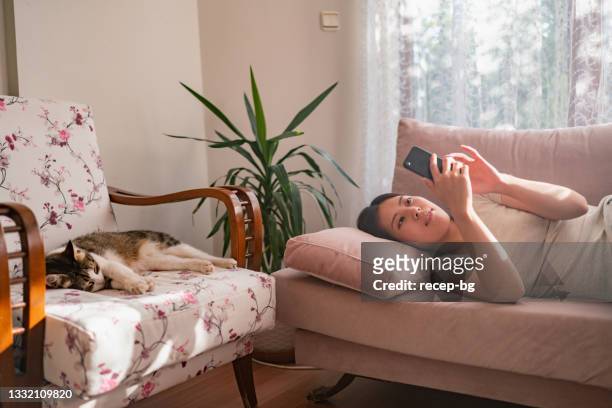 jeune femme utilisant un téléphone intelligent à la maison pendant que son chat dort à côté d’elle dans le salon à la maison - feline photos et images de collection