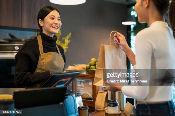 we hope you enjoy your drink of coffee. female cafe owner giving a take-out coffee to her customer at a cashier counter of a coffee shop. take-out food, point of sale system. - coffee bag stock pictures, royalty-free photos & images