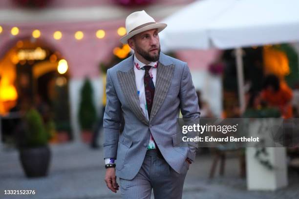 Laszlo Nagy wears a white with brown strap hat, a white with red / blue / green flower print pattern shirt, a black print pattern scarf, a gray with...