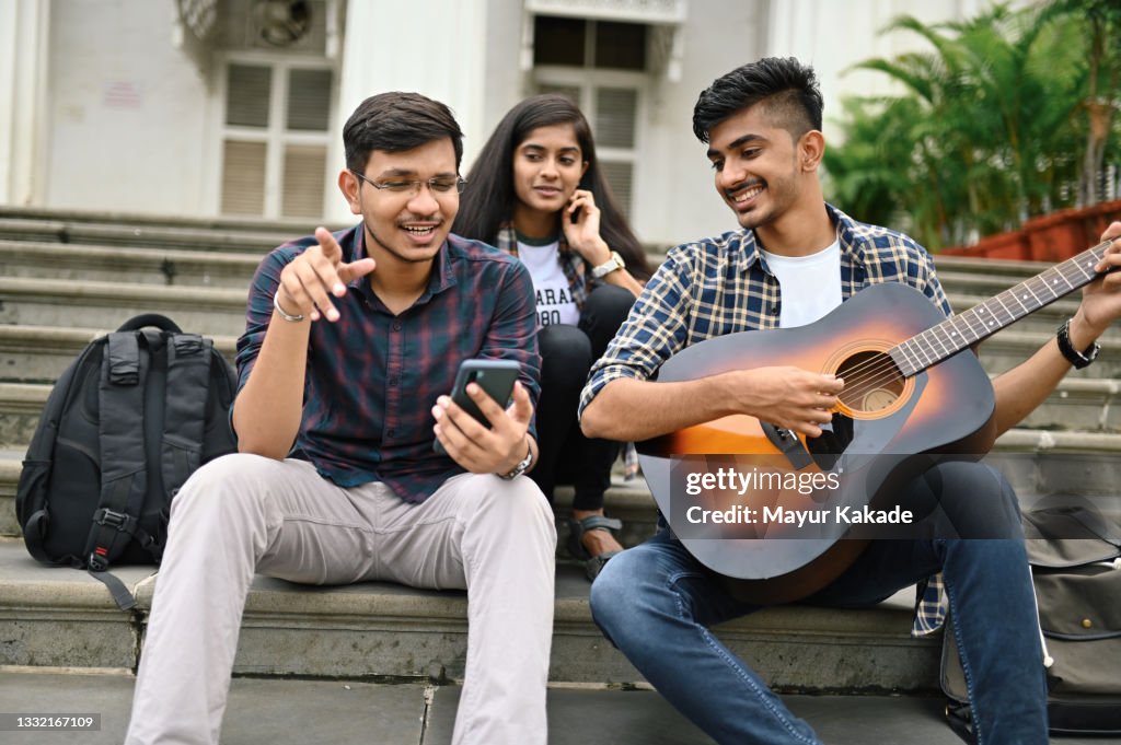 University student friends sitting on the steps of college/ university with his friend and playing guitar